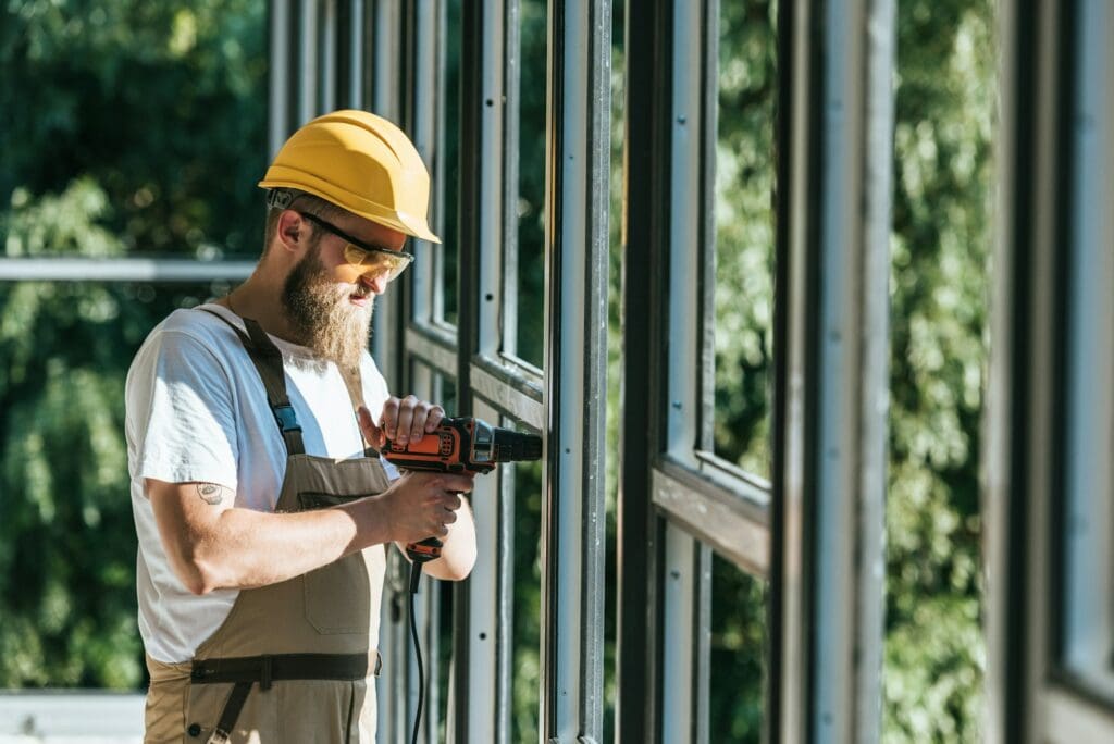 side view of builder in hardhat and protective googles installing windows by drill at construction